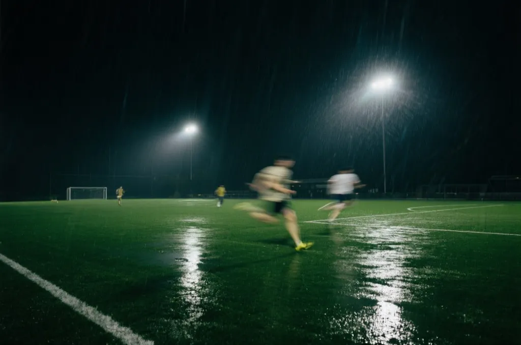 夜间球场在雨后灯光下，草皮湿滑，球员体能消耗明显